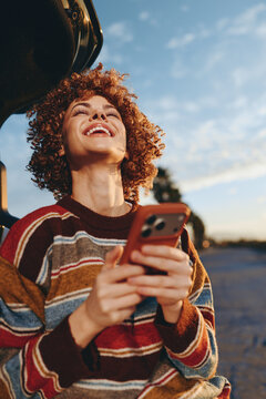 Woman with curly hair smiling outdoors, holding smartphone near road at sunset, wearing striped sweater, golden evening light, casual lifestyle, candid street portrait, relaxed mood