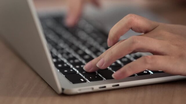 Close-up of woman's hands typing on silver laptop keyboard at wooden desk, remote work or digital content creation