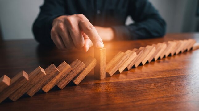 Close up of male hand stopping falling wooden dominoes on dark wooden table representing risk management and business problem solving conceptual photography with soft warm lighting and focus