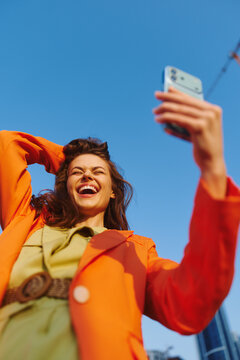 Joyful young woman in an orange jacket takes a selfie with her smartphone against a vast cloudless blue sky. Her laughing face radiates optimism. Concept of travel, freedom and happy lifestyle.
