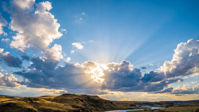 Majestic sunset beams break through dramatic fluffy white clouds over rolling golden hills and winding river