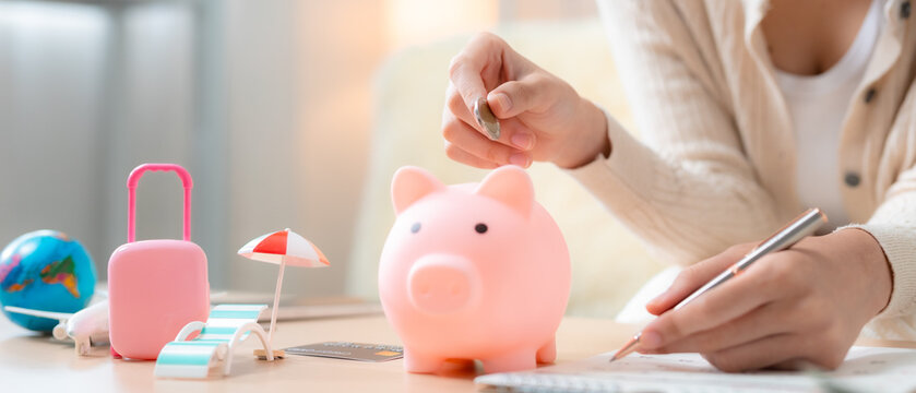 women puts coin into piggy bank at home desk while planning vacation budget with suitcase, airplane, globe and credit card, money, finance, tourism, holiday, budget. Travel savings concept.