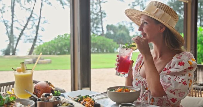 Happy woman in straw hat enjoying refreshing red cocktail at tropical beach restaurant. Smiling female tourist having lunch with healthy food and drinks on summer vacation at luxury resort.