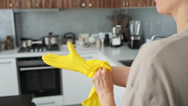 Woman wearing beige shirt pulls yellow rubber gloves onto hands in kitchen. Protective handwear supports cleaning preparation, household chores, dishwashing routines, and hygienic surface care
