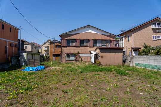 Vacant lot with weeds and an old weathered wooden house under a clear blue sky