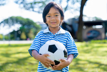 Obraz premium Portrait of happy little Asian boy holding a soccer ball looking at camera in sunny park. Active childhood and outdoor recreation concept.