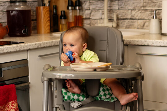 Baby Boy Bites Toy Beside Plate In Kitchen