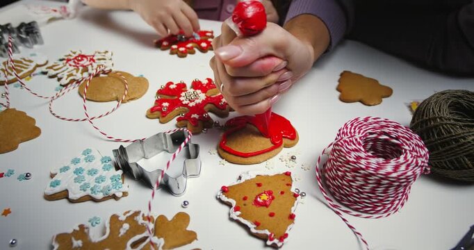 Woman hands decorating christmas gingerbread cookie with red icing on white table backdrop.