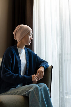 Pensive Asian woman wearing a headscarf sitting indoors and looking out the window. Cancer patient, chemotherapy recovery, mental health, and hope concept. Vertical Shot