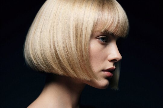 Side profile of young blonde woman with short bob haircut, bangs. Model shows elegant hairstyle, clean makeup in dark studio. Beautiful female face with smooth skin poses against black background.