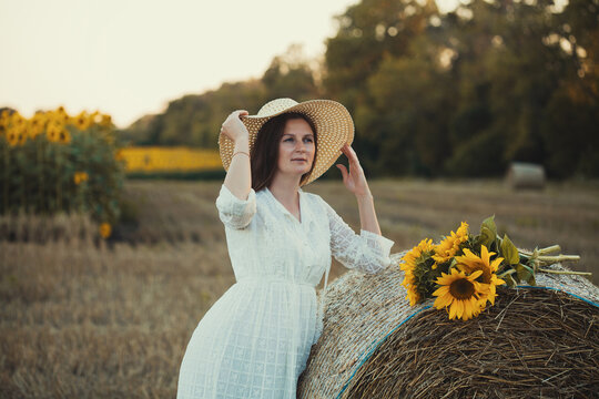 woman in a white dress holding a bouquet of wild flowers with sunflowers in a mowed field with straw bales