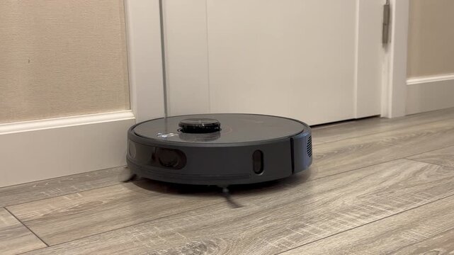 Modern black robotic vacuum cleaner sweeping a light laminate floor near an open door inside a room. Smart home appliance cleaning house doorway and driving between apartment chambers. Domestic. 