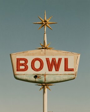 Bowling sign with weathered red letters and starburst crowns on a steel post, featuring flaking paint for nostalgia and roadside travel features
