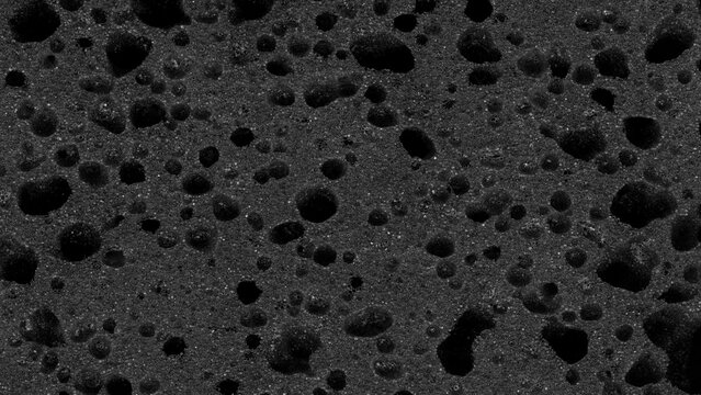 Texture of a black porous sponge (foam rubber). A macro image of the material's cellular structure, featuring numerous small voids and larger cavities.