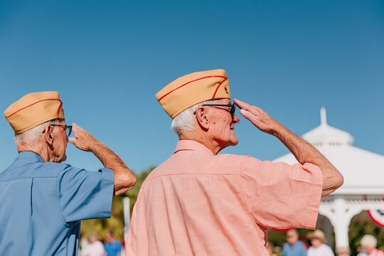 Elderly veterans in garrison caps salute during a solemn memorial ceremony, showing weathered skin and pressed shirts for remembrance and honor
