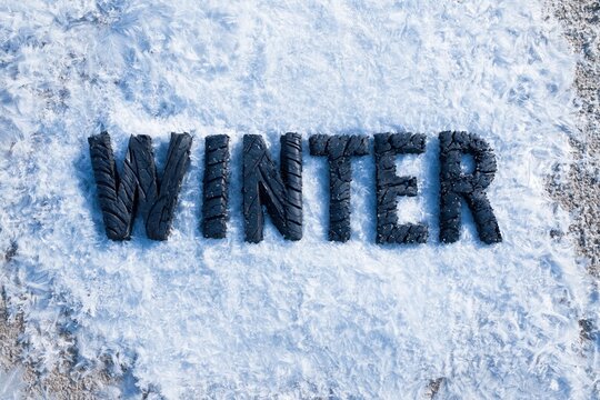 Winter letters made of black rubber tire tread resting in crystalline frost on a paved surface for automotive maintenance and seasonal care