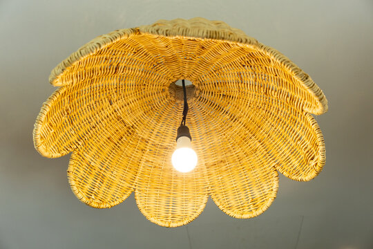 Woven rattan pendant lamp with scalloped shade and glowing bulb, viewed from below against neutral ceiling, creating warm ambient lighting and decorative interior atmosphere