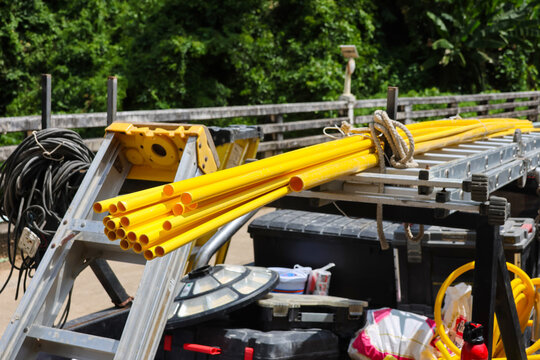 Yellow construction pipe and ladder rest on utility trailer beside equipment cases and coiled cable, creating organized outdoor work scene with lush greenery