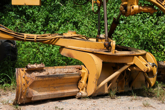 Yellow construction excavator attachment resting beside dense green vegetation, showing hydraulic arm, steel bucket, weathered metal, and heavy equipment detail in outdoor sunlight