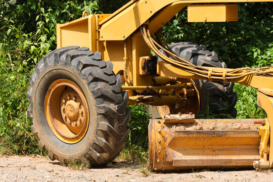 Yellow construction loader with large rugged tire and hydraulic attachment beside green foliage, showing weathered metal and outdoor earthmoving equipment in rural work setting