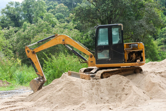 Yellow excavator parked on sand pile beside dense green forest, showing tracked construction machinery, digging equipment, hydraulic arm, bucket, and outdoor worksite scene