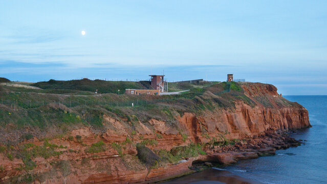 The moon rises over coastal lookout facilities on the dramatic red sandstone cliffs of Straight Point at Sandy Bay, Devon.