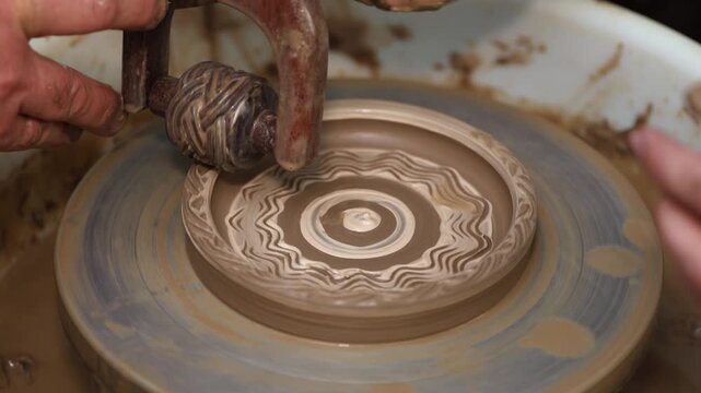 Professional artisan creating a traditional pattern on a raw clay dish using a special tool on a potter's wheel in a workshop, showcasing a detailed view of the ceramic crafting process