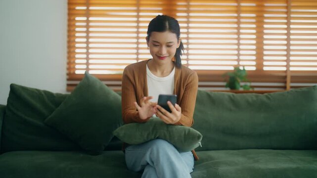 Happy young Asian woman relaxing on comfortable sofa at home, texting on smartphone. Smiling young woman using mobile phone, chatting online, shopping online from home