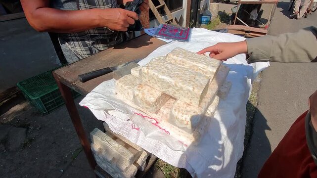 Video of Woman hands rest gently on white cloth selecting pile of tempeh. Stacked blocks sit on top of the fabric, neatly arranged. Person beside her adjusts a small blue item