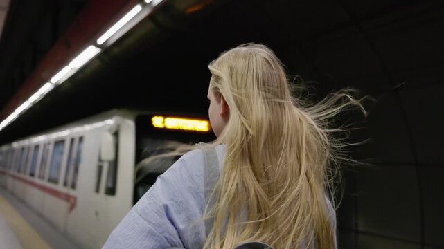 Blonde girl with long hair standing on platform as train arrives in subway station.
