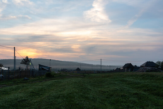 Rural village landscape with grassy meadow, scattered houses, utility poles, forested hills, and pastel sunrise sky in countryside