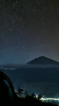 Starry night sky above misty mountain silhouette, glowing tent rests on high ground, distant lights twinkle beneath endless sea of stars