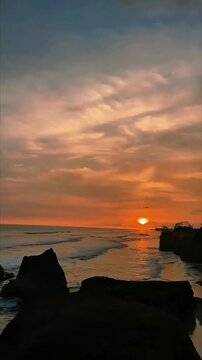 Golden sunset spills over calm ocean, waves gently kiss rocky shoreline, warm clouds drift across glowing sky in perfect harmony