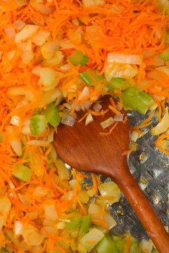 A close up top view shot of a traditional mirepoix vegetable mixture being stirred with a wooden spoon in a shiny metal pan during cooking.
