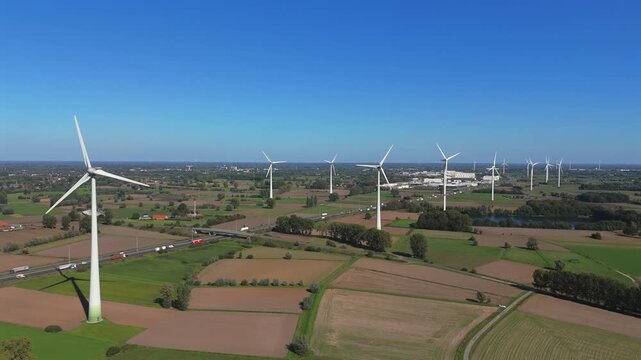 Drone footage starting from a wider distance and moving forward toward multiple wind turbines in the Belgian countryside, with a highway and additional turbines