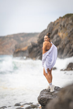 Woman stands on rock by the ocean, looking back while waves crash on the beach in a coastal area