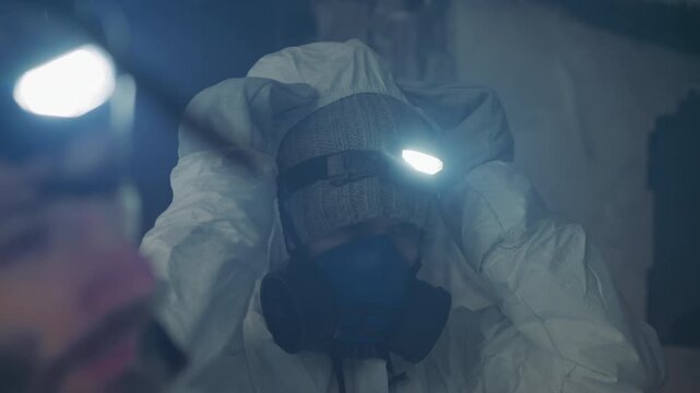 Technician in protective suit adjusting respirator and headlamp in dim ruined house, knitted hat visible, gloved hands securing strap, dust and plaster falling, tense preparation for sample