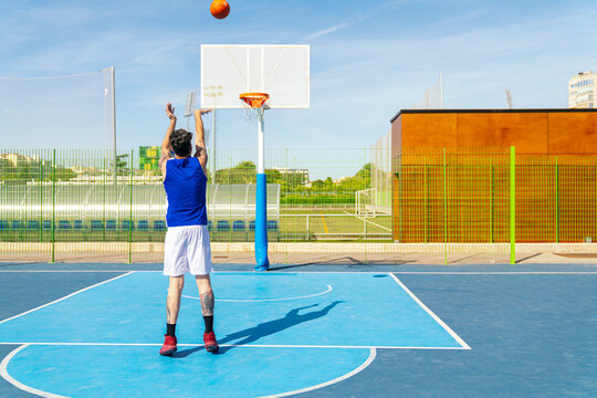 Athlete wearing blue jersey and white shorts shooting a basketball towards the hoop on a vibrant blue outdoor court