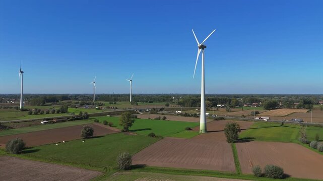 Drone footage starting from a wider distance and moving forward toward multiple wind turbines in the Belgian countryside, with a highway and additional turbines visible