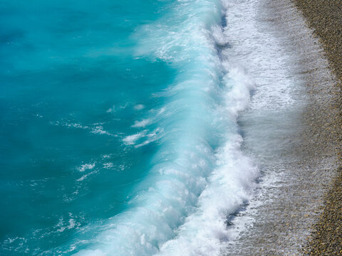 Big storm waves crashing on a pebble beach with azure water. Summer sea coastline during powerful ocean weather.