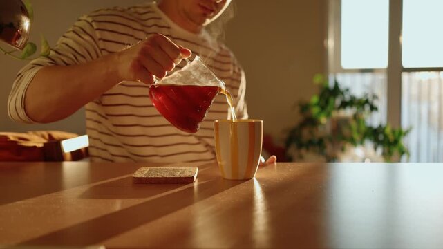 Daily morning routine of a man pouring hot, steaming coffee from a glass pot into a cup, sitting at a wooden table illuminated by the warm light of the rising sun from a nearby window
