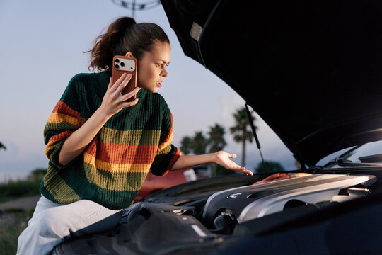 Young woman calling for help on smartphone near car with open hood. Checking broken engine at dusk. Concept of car trouble, roadside emergency and breakdown assistance.