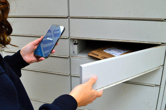 Young woman holding a smartphone displaying a Vinted app while opening an automated parcel locker to collect her online shopping delivery outdoors