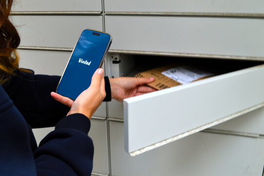 Woman holding a smartphone displaying a secondhand marketplace app while retrieving a cardboard package from an automated parcel locker outdoors