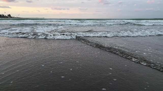 Static ground-level slow motion view of ocean waves reaching a sandy shore with an open sky stretching across the distant horizon, creating a calm coastal atmosphere.