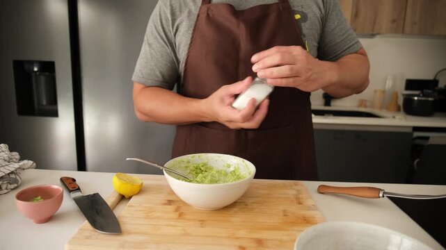 Man wearing an apron squeezing lemon and adding salt to a bowl of fresh homemade guacamole sauce
