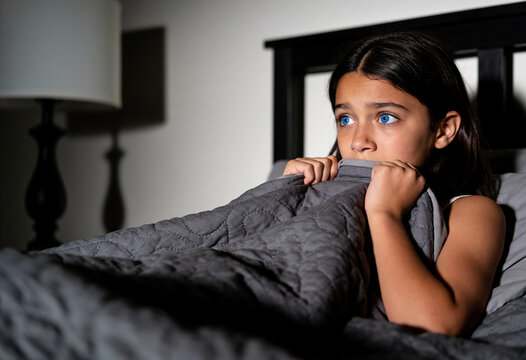 Scared young girl hiding under grey blanket in dark bedroom looking fearful with wide blue eyes at night shadows