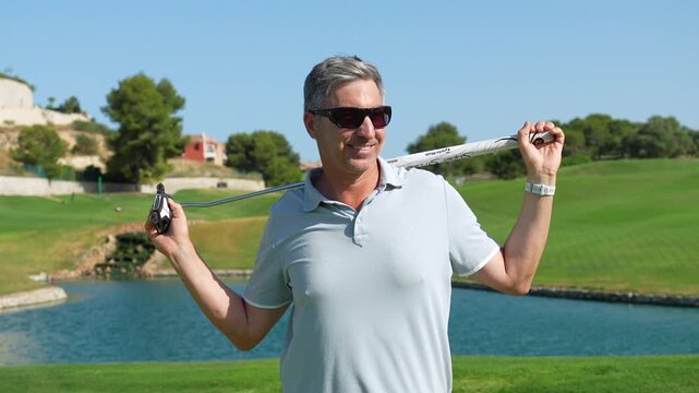 Successful mature golfer smiling confidently at the camera while holding a putter behind his shoulders, standing on a beautiful green course with a water hazard in the background on a sunny day