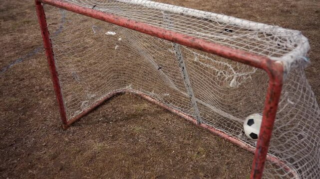 worn goal with lone ball on dry field. frayed net and bent red frame on parched grass, solitary ball resting near goalmouth, muted earth tones and scattered debris, abandoned practice atmosphere