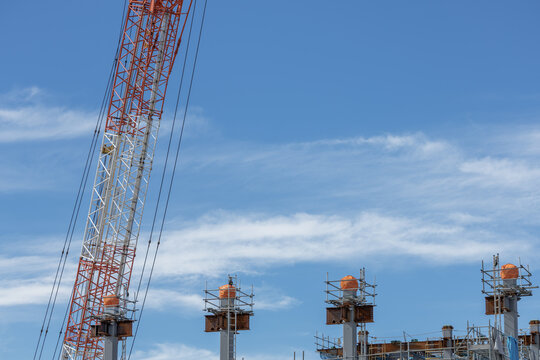 Lattice boom crane jib towering over orange-capped steel column framework of a building structure at a construction site set against a bright blue sky with wispy clouds.